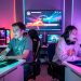 Scène dynamique d’un marathon de streaming gaming caritatif avec un homme et une femme devant des écrans, éclairés par des lumières cyan, magenta et violettes, avec un indicateur de dons lumineux sans inscription.